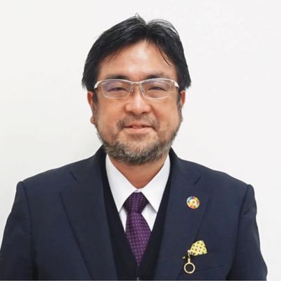 黒いスーツにマフラーをまいて帽子をかぶっている。髭を生やし、メガネをかけている40代の男性が正面を向いて微笑んでいる。